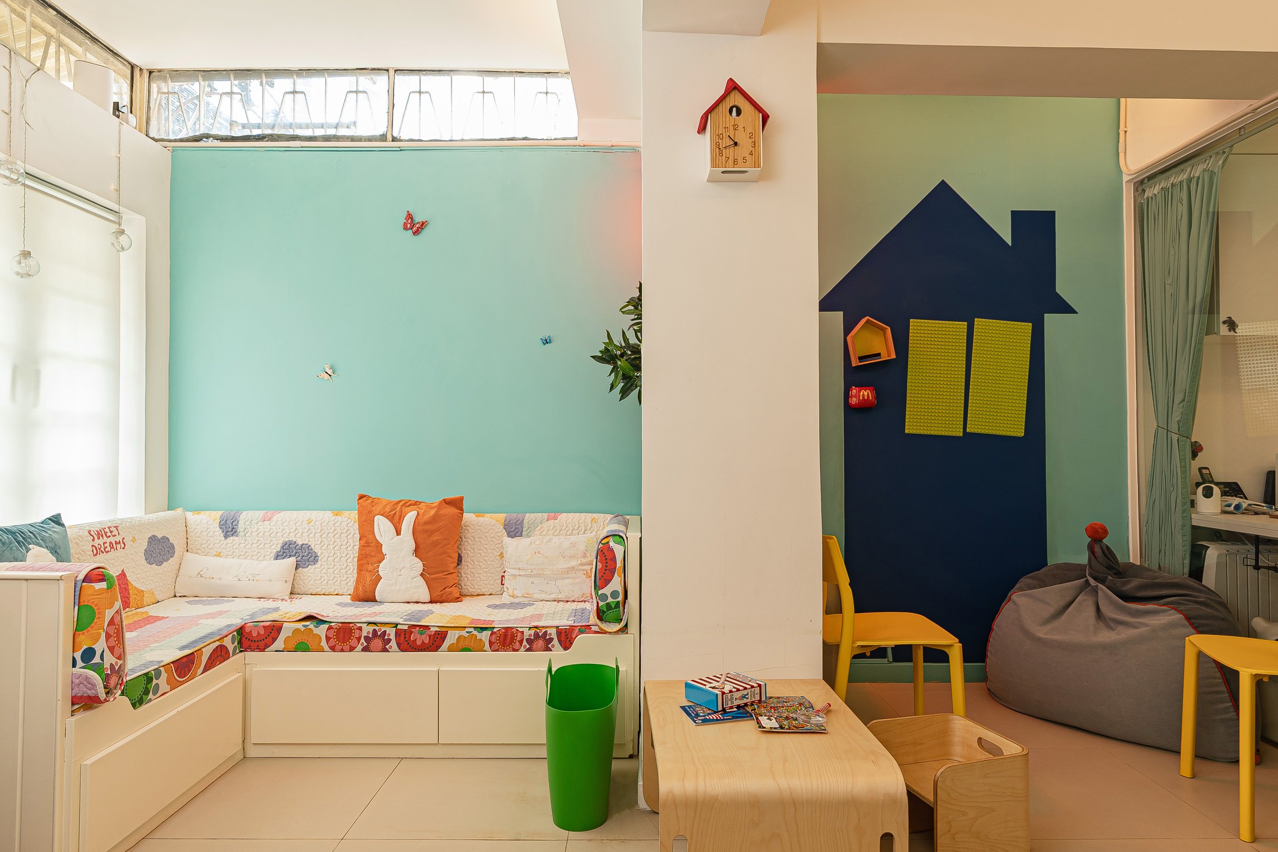 A brightly lit, child-friendly room divided into two sections. On the left, there's a cozy corner with a white L-shaped bench adorned with colorful, patterned cushions and pillows — including one with a bunny design and another that says “Sweet Dreams.” The wall is painted a soft teal, decorated with 3D butterfly accents. On the right side, a small play area features a bean bag, yellow chairs, and a wall painted with a blue house silhouette and mounted yellow pegboards. A light wooden table with toys and books sits in the foreground, and a red-roofed wall clock is mounted on a dividing pillar.