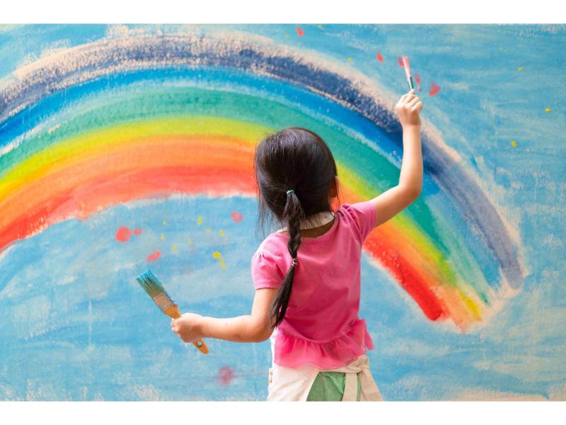 a lovely girl wearing a pink dress drawing a rainbow on the wall