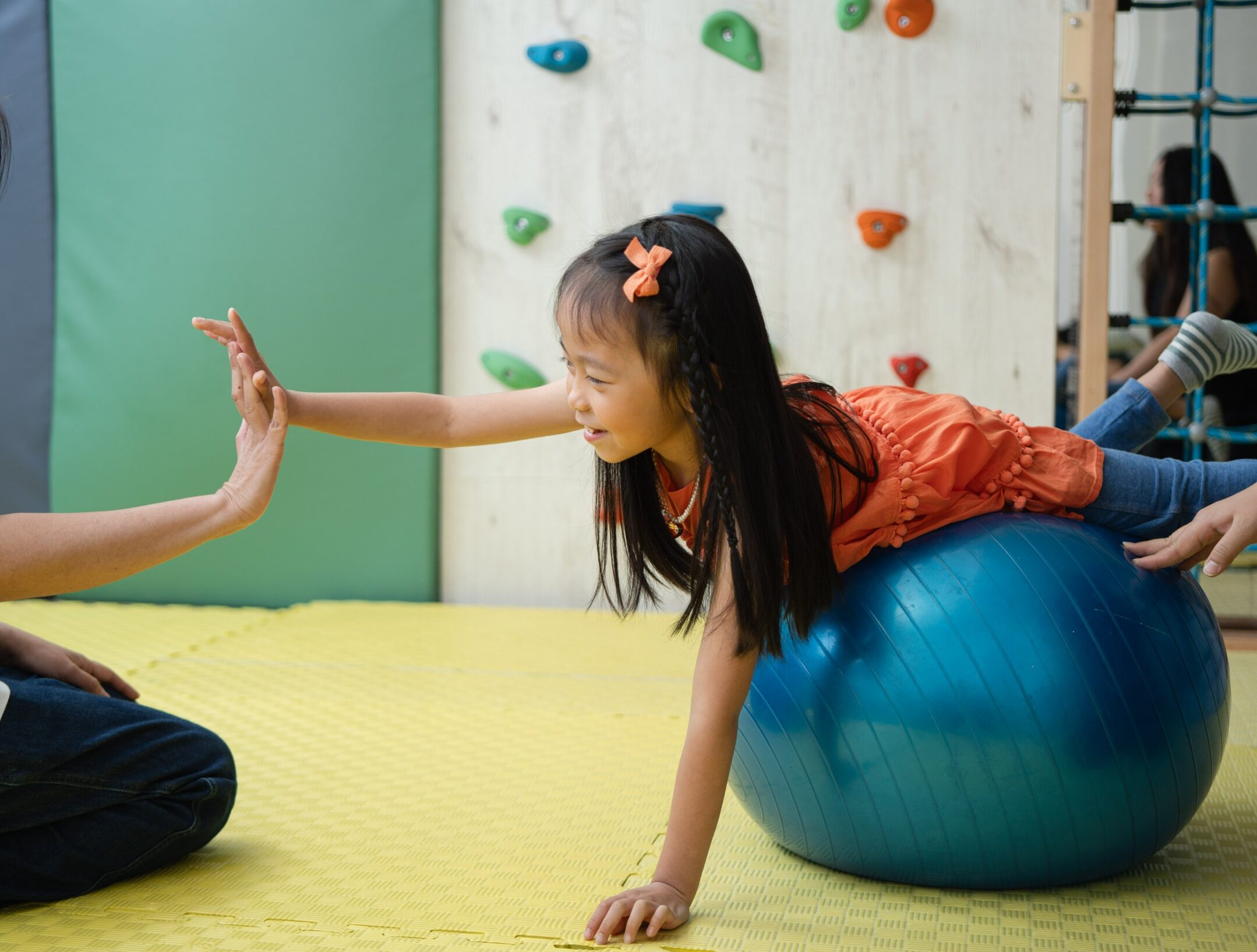 A sweet 5 year old girl balancing on a blue yoga ball and trying to high fice an Occupational therapist in a fully equipped and padded OT room in Sprout in Motion Wong Chuk Hang Centre.