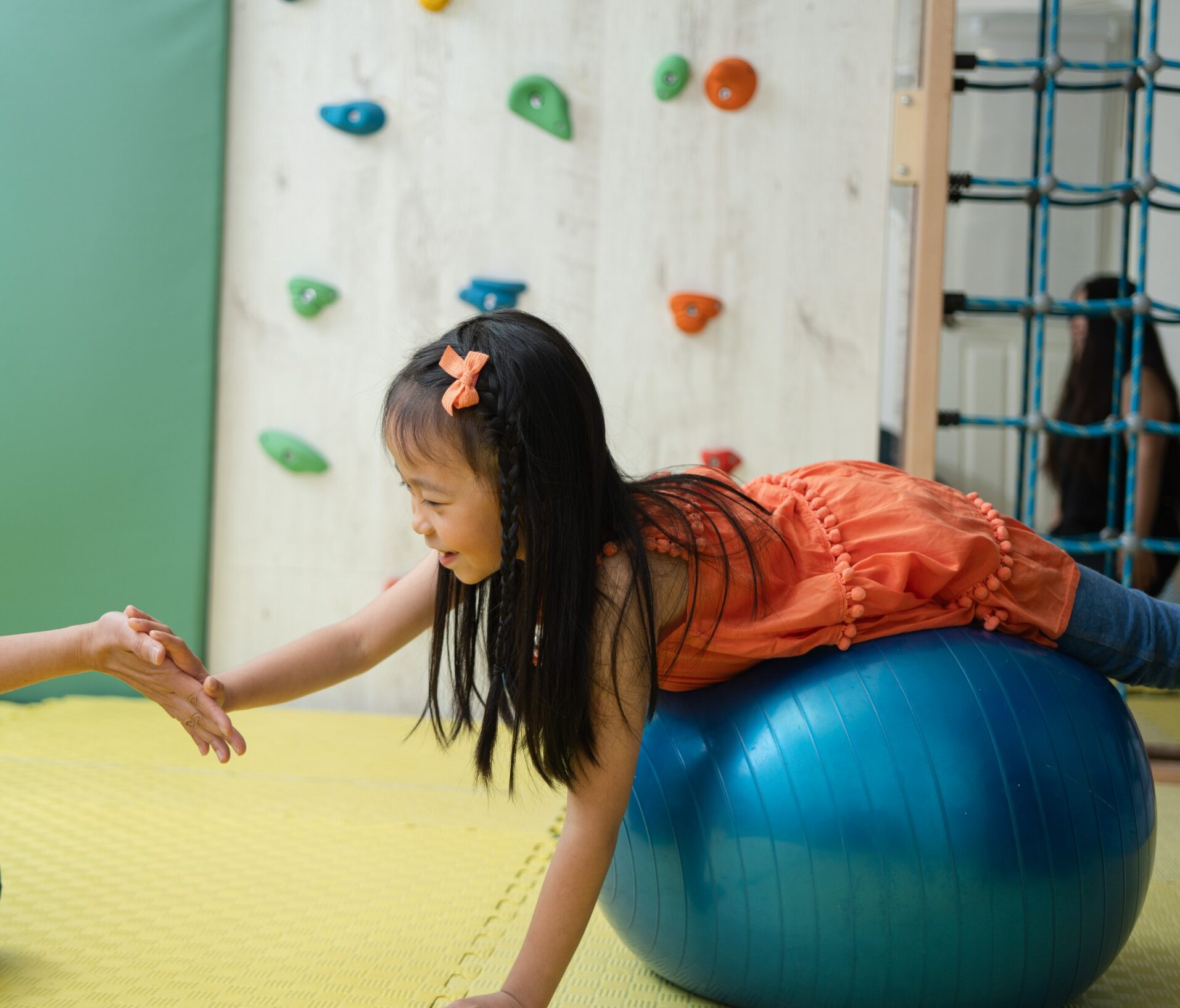 A sweet 5 year old girl balancing on a blue yoga ball and trying to high fice an Occupational therapist in a fully equipped and padded OT room in Sprout in Motion Wong Chuk Hang Centre.