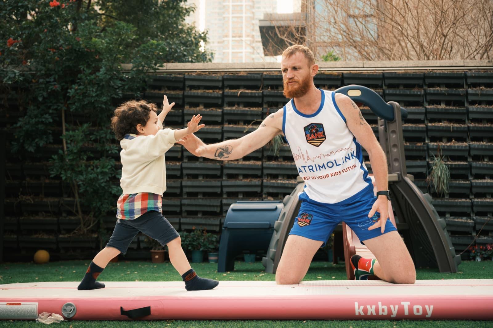JC Richards guiding a child stepping across a gymnastics mat with arms raised.