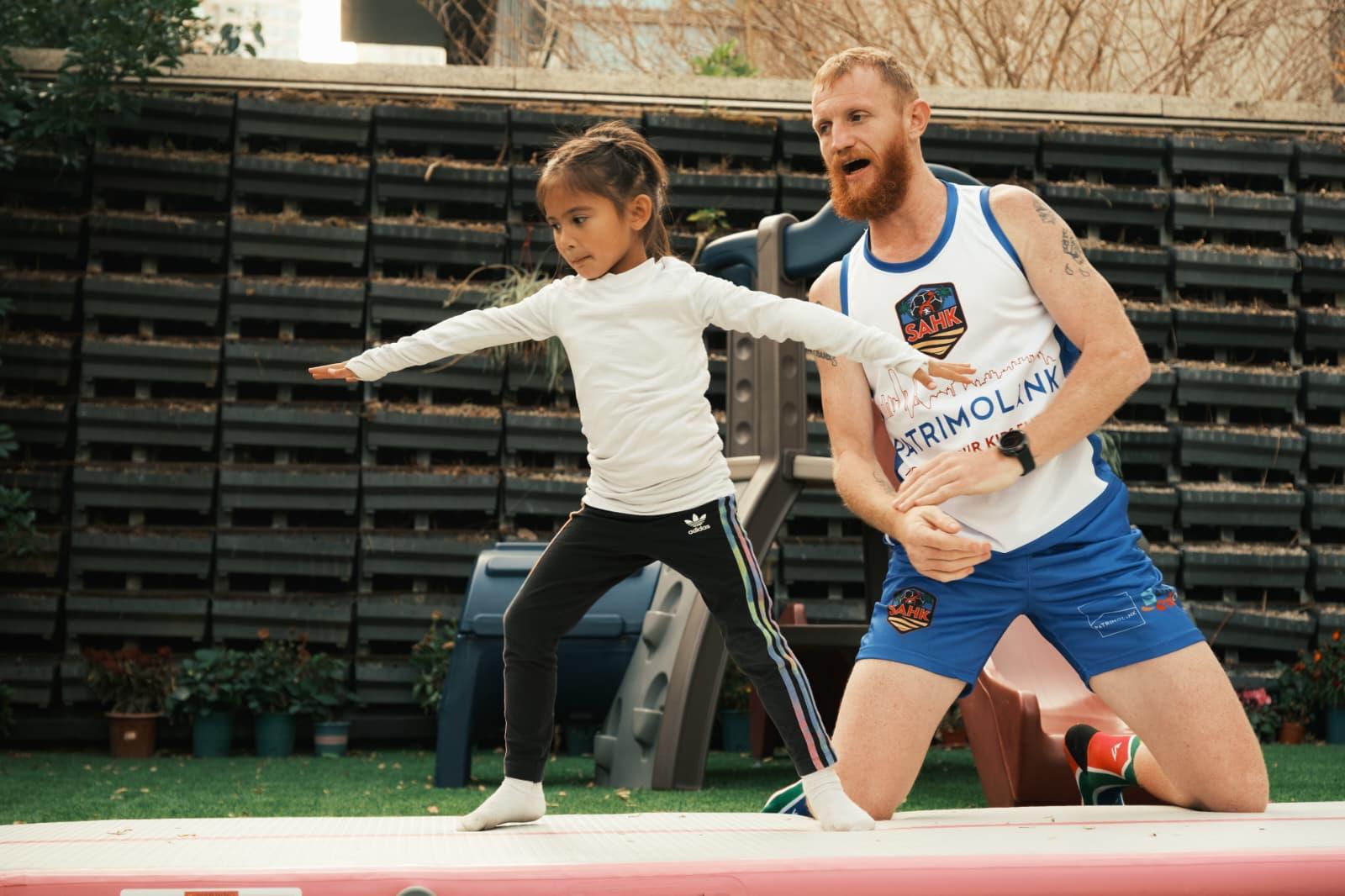 JC Richards kneeling beside a child practising a wide stance with arms outstretched on an outdoor gymnastics mat.