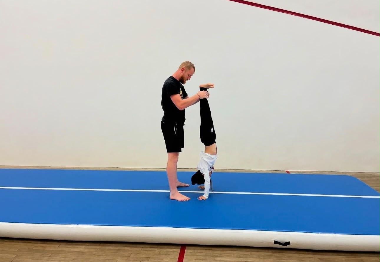JC Richards supporting a child’s lower legs during a handstand on a blue gymnastics mat.