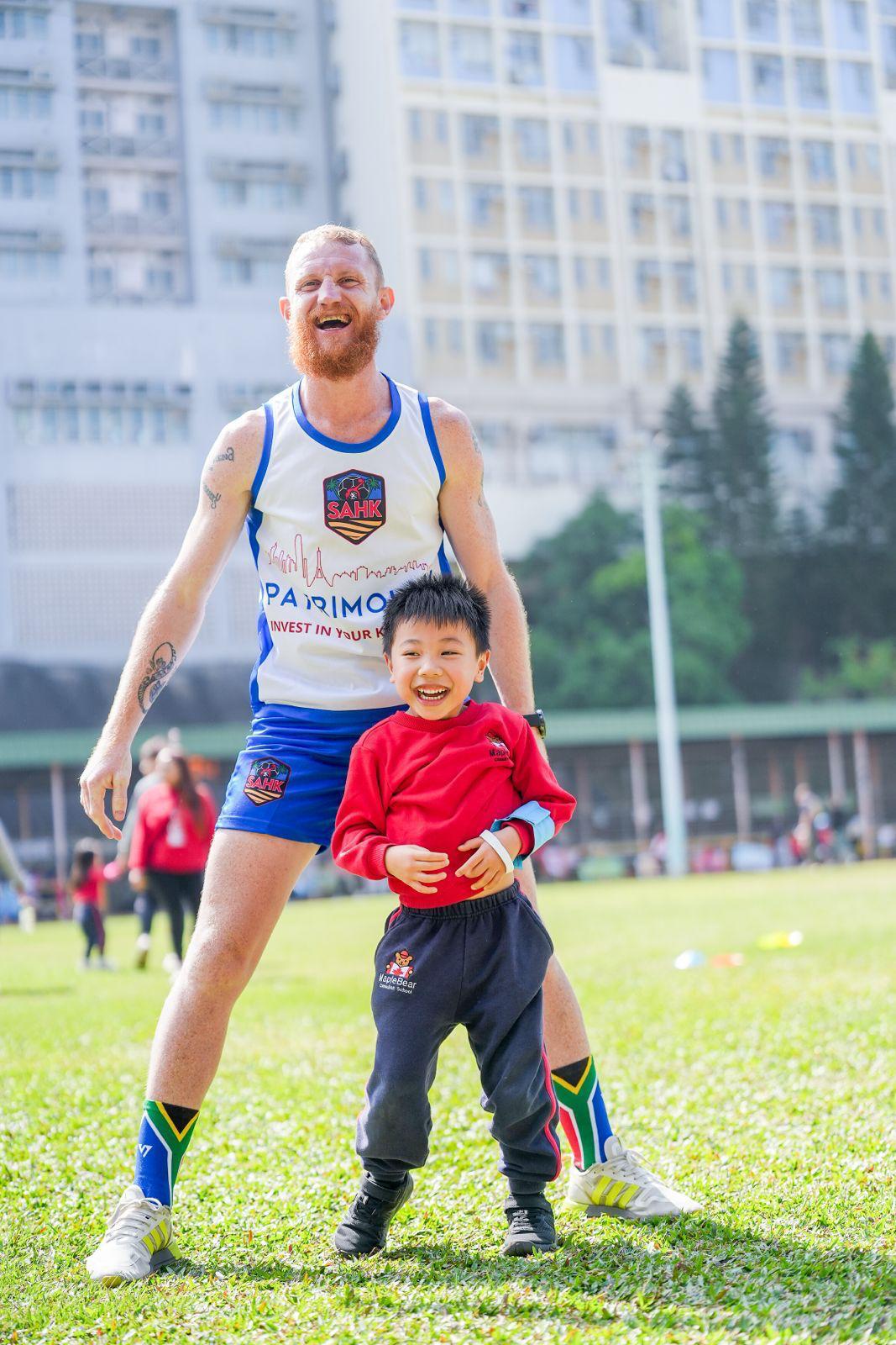 Gymnastics coach Juan-Claude (JC) Richards smiling with a child on a grass field.